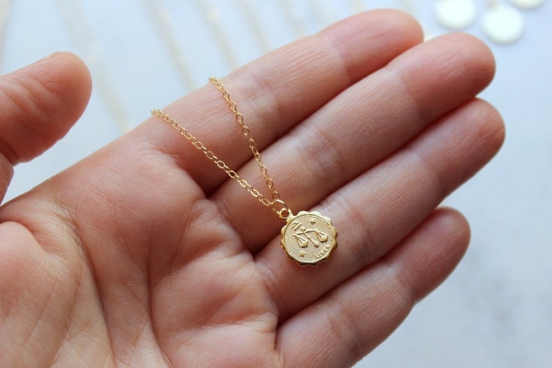 May include: Gold chain necklace with a small, round pendant featuring the zodiac symbol for Libra.