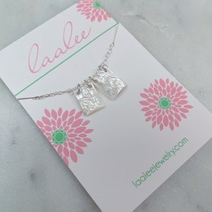 May include: A silver necklace with two rectangular pendants featuring embossed images. The necklace is displayed on a white card with pink floral designs and the text "laalee" and "laaleejewelry.com".