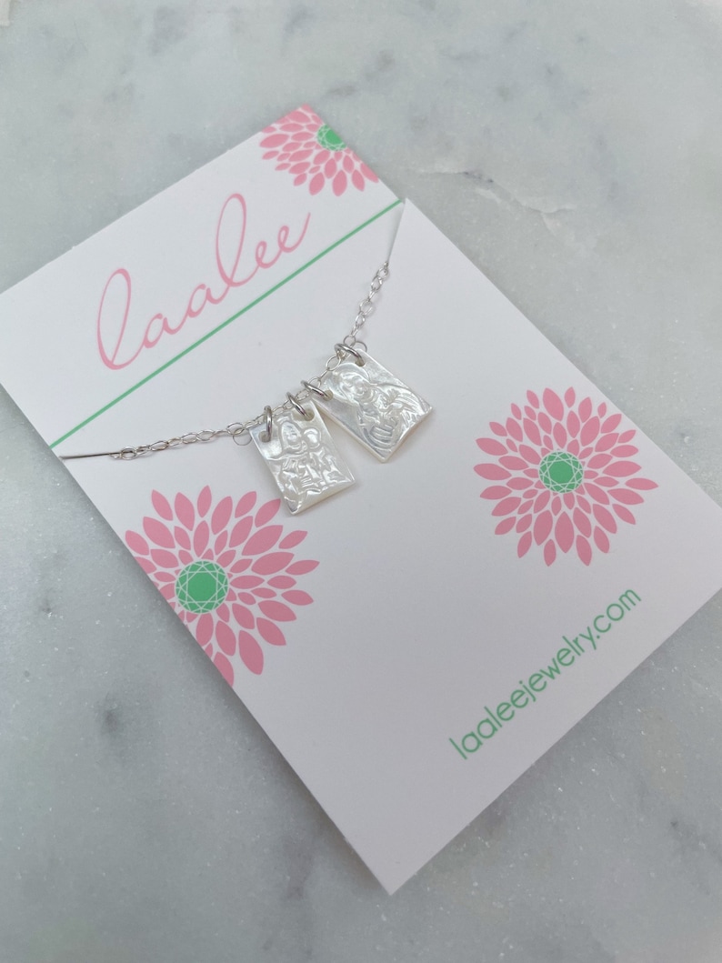 May include: A silver necklace with two rectangular pendants featuring embossed images. The necklace is displayed on a white card with pink floral designs and the text "laalee" and "laaleejewelry.com".