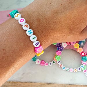 May include: A close-up of two colorful beaded bracelets. One bracelet spells "SOPHIE" with white letter beads and flower accents. The other bracelet spells "CHARLOTTE" and "MYA" with white letter beads and flower accents. The bracelets are on a light background.