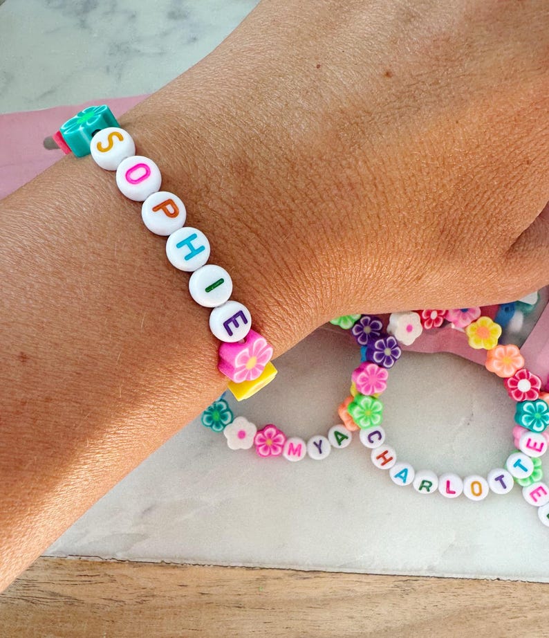 May include: A close-up of two colorful beaded bracelets. One bracelet spells "SOPHIE" with white letter beads and flower accents. The other bracelet spells "CHARLOTTE" and "MYA" with white letter beads and flower accents. The bracelets are on a light background.