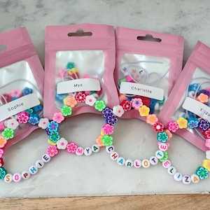 May include: Four pink pouches containing bracelet-making kits. Each kit includes colorful beads and flower-shaped accents. The bracelets spell out names: Sophie, Mya, Charlotte, and Eliza. The bracelets are adorned with flower beads.