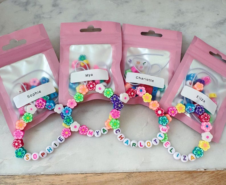 May include: Four pink pouches containing bracelet-making kits. Each kit includes colorful beads and flower-shaped accents. The bracelets spell out names: Sophie, Mya, Charlotte, and Eliza. The bracelets are adorned with flower beads.