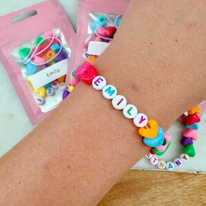 May include: A colorful beaded bracelet with the name "EMILY" spelled out in white letters. The bracelet is made with heart-shaped beads in various colors, including pink, yellow, blue, green, and orange. The bracelet is on a person's wrist, and there are two bags of beads in the background.