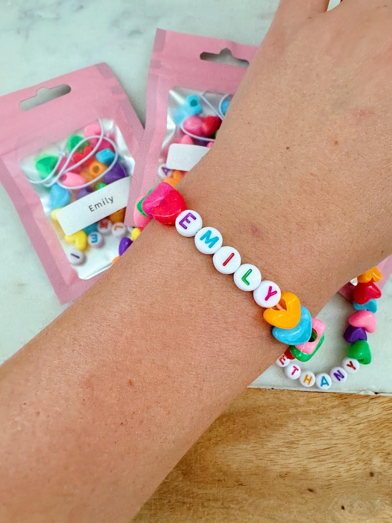 May include: A colorful beaded bracelet with the name "EMILY" spelled out in white letters. The bracelet is made with heart-shaped beads in various colors, including pink, yellow, blue, green, and orange. The bracelet is on a person's wrist, and there are two bags of beads in the background.