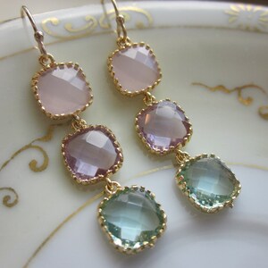 May include: A pair of gold dangle earrings with three faceted gemstone drops. The top drop is light pink, the middle drop is a darker pink, and the bottom drop is a light blue-green.