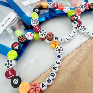 May include: A close-up of three colorful beaded bracelets with the names "Andrew", "William", and "John Luke" spelled out in white beads. The beads are decorated with various sports-themed designs, including basketballs, baseballs, footballs, and soccer balls.