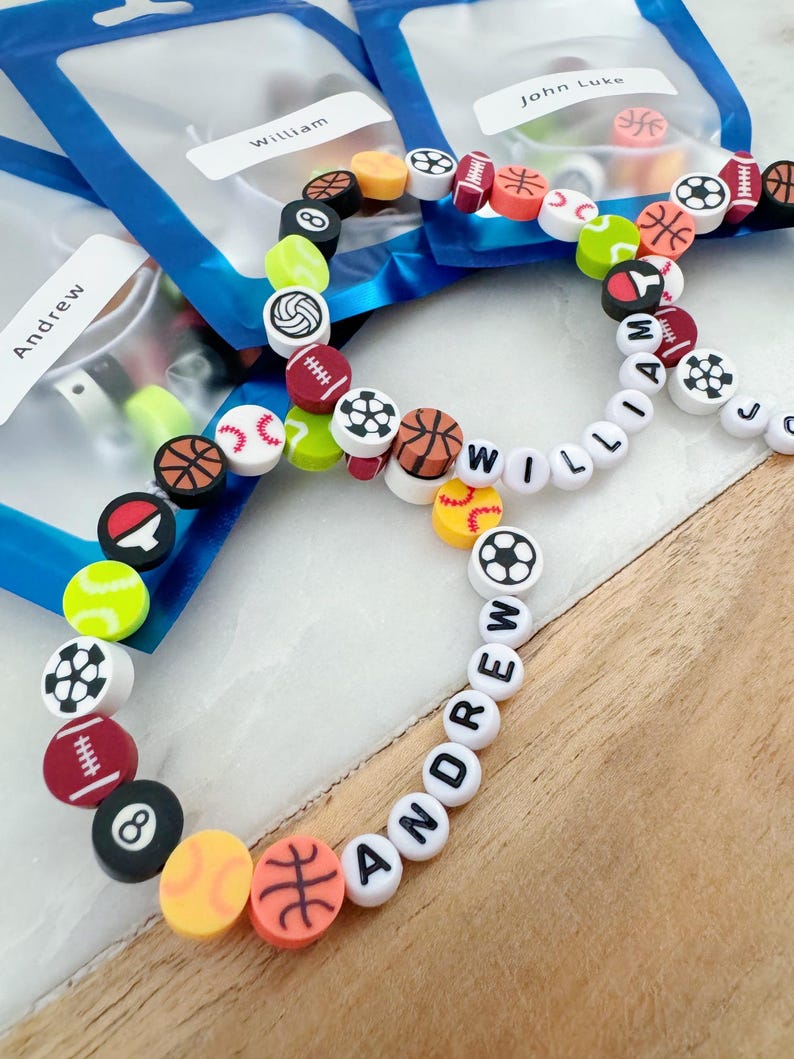 May include: A close-up of three colorful beaded bracelets with the names "Andrew", "William", and "John Luke" spelled out in white beads. The beads are decorated with various sports-themed designs, including basketballs, baseballs, footballs, and soccer balls.