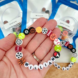 May include: A white beaded bracelet with the name "William" spelled out in black letters. The bracelet is decorated with colorful sports-themed beads, including basketballs, footballs, soccer balls, baseballs, and tennis balls.