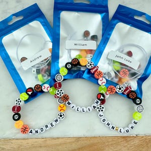 May include: Three clear plastic bags with white string and colorful sports-themed beads. The bags are labeled "Andrew", "William", and "John Luke". Three bracelets are laid out on a white surface, each with the name "Andrew", "William", and "John Luke" spelled out in white beads.