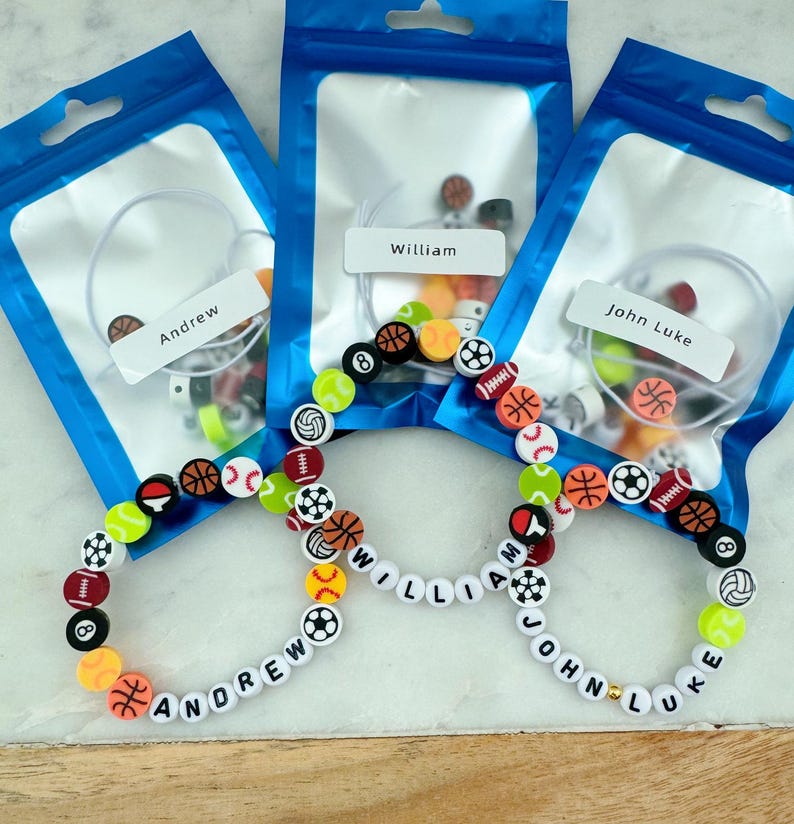 May include: Three clear plastic bags with white string and colorful sports-themed beads. The bags are labeled "Andrew", "William", and "John Luke". Three bracelets are laid out on a white surface, each with the name "Andrew", "William", and "John Luke" spelled out in white beads.
