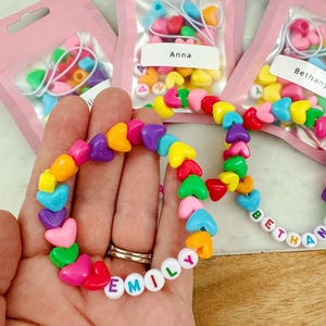May include: Three small pink bags with clear plastic windows containing colorful heart-shaped beads and letter beads. The bags are labeled "Anna", "Bethany", and "Emily". A hand is holding a bracelet made with the beads, spelling out the name "Emily".