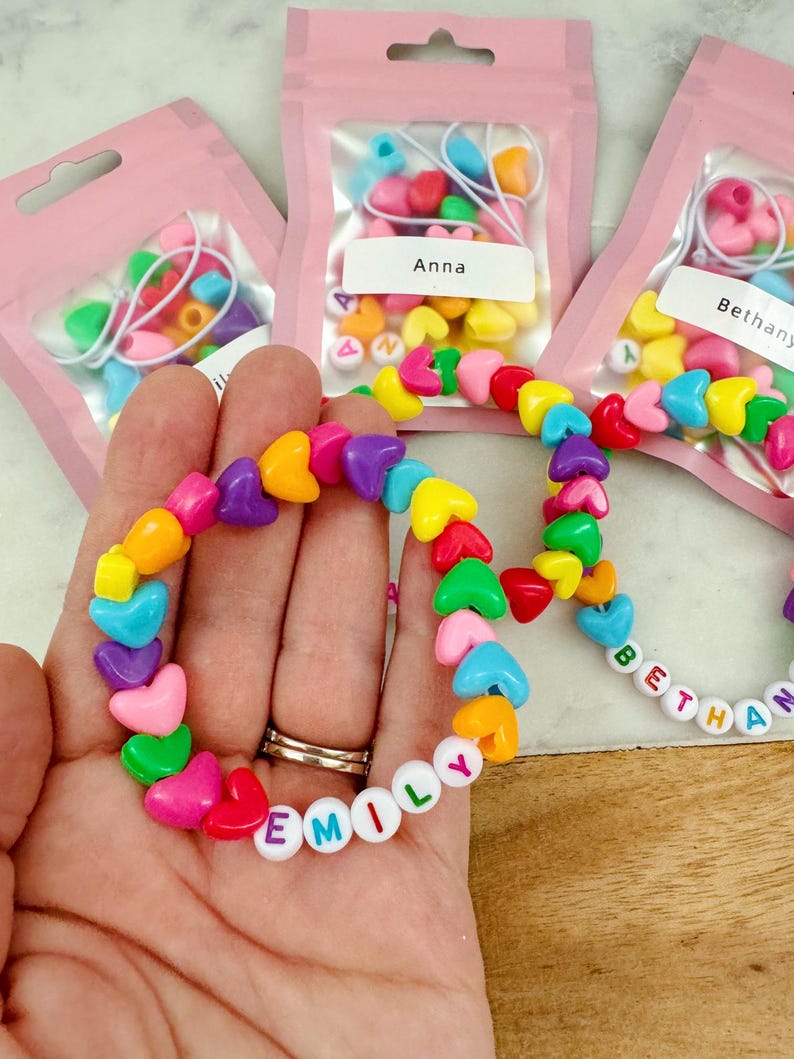 May include: Three small pink bags with clear plastic windows containing colorful heart-shaped beads and letter beads. The bags are labeled "Anna", "Bethany", and "Emily". A hand is holding a bracelet made with the beads, spelling out the name "Emily".
