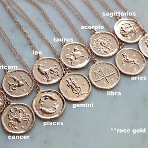 May include: Twelve gold-toned zodiac necklaces with pendants featuring astrological symbols. The necklaces are arranged in a circle on a white surface. The text "rose gold version" is visible at the bottom of the image.