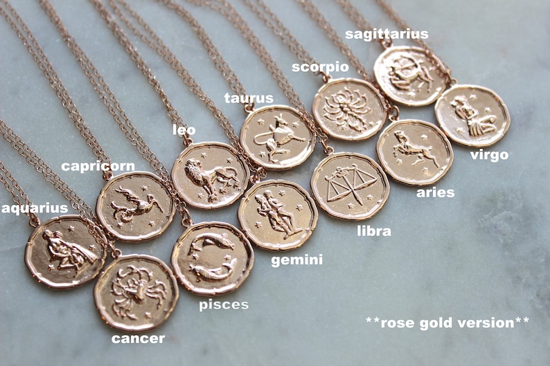 May include: Twelve gold-toned zodiac necklaces with pendants featuring astrological symbols. The necklaces are arranged in a circle on a white surface. The text "rose gold version" is visible at the bottom of the image.