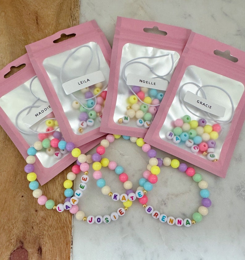 May include: Five pink bags with white string and colorful beads inside. The bags are labeled with the names Maddie, Leila, Noelle, Gracie, and the names Laalee, Josie, Kate, and Brenna are spelled out with beads on the table.