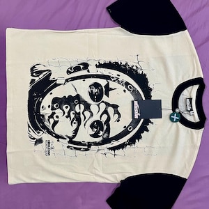 Pode incluir: Camiseta creme com mangas pretas e gola redonda. A frente apresenta um grande gráfico preto desgastado de um design semelhante a um crânio dentro de uma moldura circular. As mangas têm um logotipo branco. A camisa tem uma etiqueta.