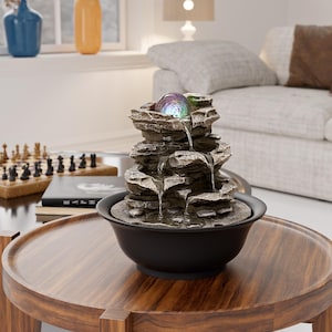 May include: An indoor water feature with a layered rock design. The fountain has a glowing, multi-coloured orb at the top and water cascading into a black bowl. The fountain sits on a round, wooden table.