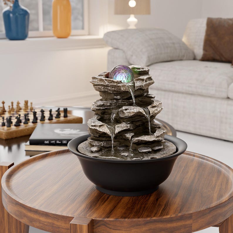 May include: An indoor water feature with a layered rock design. The fountain has a glowing, multi-coloured orb at the top and water cascading into a black bowl. The fountain sits on a round, wooden table.