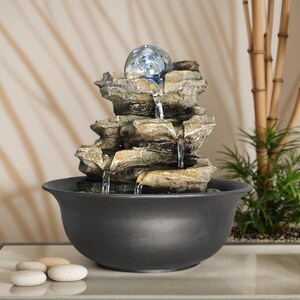 May include: An indoor water feature with a layered rock structure and a clear glass sphere. Water flows down into a black ceramic bowl. The fountain is designed to create a relaxing atmosphere in a home or office.