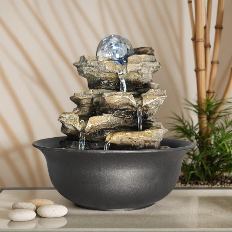 May include: An indoor water feature with a layered rock structure and a clear glass sphere. Water flows down into a black ceramic bowl. The fountain is designed to create a relaxing atmosphere in a home or office.