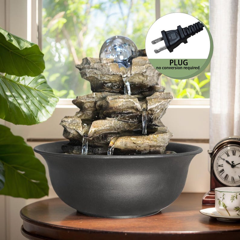 May include: An indoor water feature with a layered rock design and a clear glass sphere. Water flows into a black bowl. The fountain has a standard US plug, labelled "no conversion required". The fountain is suitable for use in the UK.