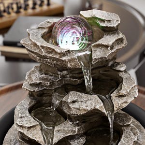 May include: An indoor water feature with a layered rock design. A clear glass orb at the top emits colourful light. Water cascades down the rock structure into a lower basin, creating a tranquil atmosphere.