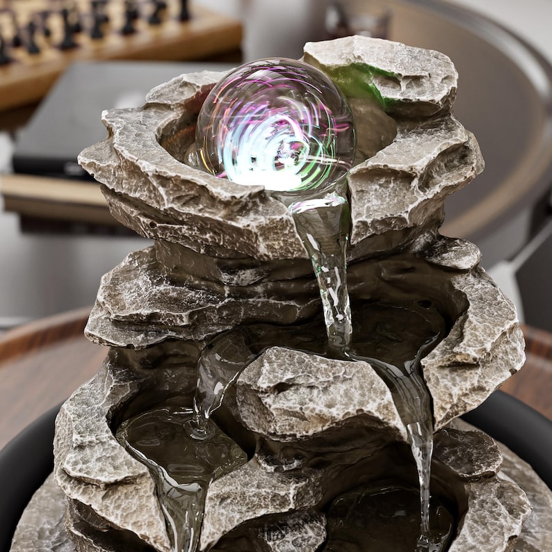 May include: An indoor water feature with a layered rock design. A clear glass orb at the top emits colourful light. Water cascades down the rock structure into a lower basin, creating a tranquil atmosphere.