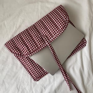 以下が含まれることがあります： 赤と白のギンガムチェックのノートパソコンスリーブ。紐で留められています。銀色のノートパソコンが一部見えています。生地はキルティング加工が施されています。スリーブは白いシーツの上に置かれています。