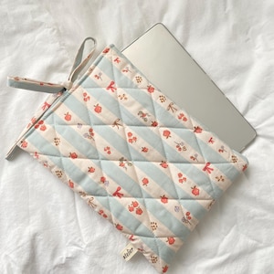 May include: A quilted laptop sleeve featuring a light blue and white striped design, decorated with small red strawberries and bows. It has a matching handle and the word "atelier" is printed on a small tag.