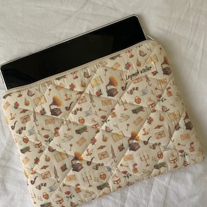 Puede incluir: Funda acolchada para portátil de color crema con un estampado repetido de ilustraciones vintage, incluyendo gramófonos, rosas y llaves. La funda tiene cierre de cremallera y el texto "Leymeh atelier". Diseñada para proteger un portátil o tableta.