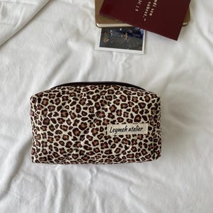Puede incluir: Un neceser rectangular con un estampado de leopardo en marrón y beige. La bolsa tiene una cremallera negra y una etiqueta blanca que dice "Leymeh atelier". La bolsa está sobre una superficie blanca.