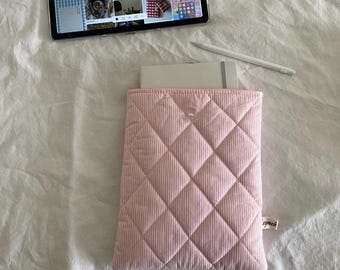 May include: A light pink corduroy tablet sleeve with a quilted diamond pattern. A white notebook is partially visible inside the sleeve. A tablet and stylus are also in the image, resting on a white surface.