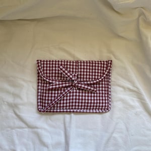 May include: A rectangular fabric pouch featuring a dark red and white gingham check pattern. The pouch has a rounded flap closure secured with two thin fabric ties that are knotted together at the center of the front panel.