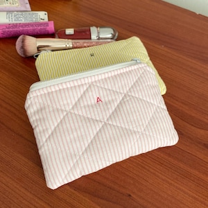 May include: A pink and white striped quilted cosmetic bag with a white zipper and the letter "A" on the front. A yellow and white striped cosmetic bag and other makeup items are in the background.