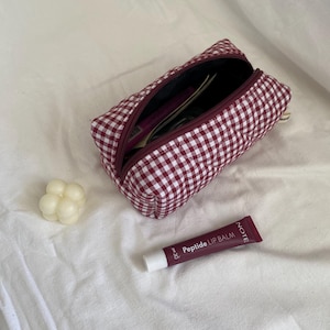 Pode incluir: Uma bolsa acolchoada com fecho de correr em xadrez vichy bordô e branco com interior escuro. Ao lado da bolsa encontra-se uma pequena vela de bolhas cor de creme e um tubo bordô de bálsamo labial Note Peptide com texto branco.