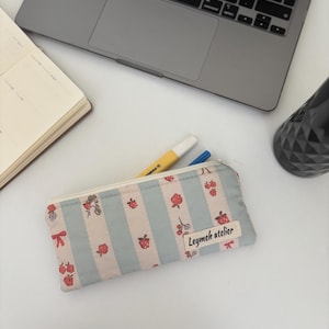 Peut inclure: Une trousse à crayons à rayures bleues et blanches, ornée de fraises et de nœuds rouges. La trousse a une fermeture éclair et une petite étiquette avec l'inscription "Leymeh atelier".