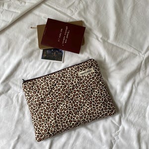 May include: A quilted laptop sleeve with a leopard print pattern in brown and tan. The sleeve has a zipper closure and a small label that reads "Leymek atelier". A burgundy book and a small photo are also visible.