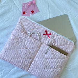 Op de afbeelding: Roze en wit gestreepte gewatteerde laptoptas met een witte rits en een rode strik. De tas heeft een vak met een notitieboekje en een wit snoer. Twee roze kaarten met hartjes en de tekst "Thank you" zijn ook zichtbaar.