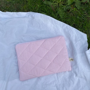 Könnte beinhalten: Eine rosa-weiß gestreifte, gesteppte Laptoptasche mit Reißverschluss. Die Hülle hat ein Rautenmuster und ein kleines Etikett. Sie liegt auf einem weißen Laken.