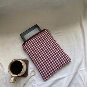 May include: A rectangular fabric sleeve featuring a dark red and white gingham check pattern, containing a black e-reader. Beside the sleeve is a speckled ceramic mug filled with dark coffee on a square wooden coaster and a small white spoon.