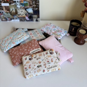 Puede incluir: Una colección de bolsas de tela acolchadas en varios patrones y colores. Los diseños incluyen estampados florales, a rayas y estampados. Cada bolsa tiene un cierre de cremallera y una etiqueta que dice "Leymeh atelier".