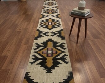 May include: A long, rectangular runner rug with a geometric pattern in black, gold, and cream. The rug has a fringed edge and is placed on a hardwood floor. The design features repeating diamond and arrow shapes.