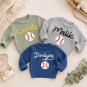 May include: Three knitted sweaters in green, grey, and blue. Each sweater features a baseball graphic and a name: "Saints", "Malik", and "Dodgers". The sweaters have ribbed cuffs and a crew neck.