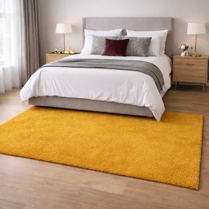 May include: A mustard yellow rectangular shag rug in a bedroom setting. The rug is placed in front of a bed with white bedding, gray pillows, and a gray headboard. Two wooden nightstands with lamps flank the bed.