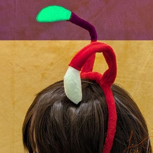 May include: A red headband with a plush bud tip, approximately 7 inches in length. The bud tip is green, with a purple stem. The headband has a white accent. The text "Small Bud Tip - 7 inches" is visible.