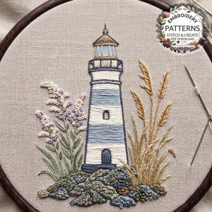 Könnte beinhalten: Ein Stickrahmen mit einem Leuchtturmmotiv auf beigem Stoff. Der Leuchtturm hat horizontale blaue und weiße Streifen, ein dunkles Dach und eine kleine Tür. Er ist von violetten Blumen und goldenen Weizenähren auf felsigem Untergrund umgeben.