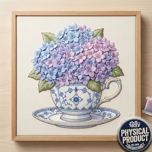 Puede incluir: Obra de arte bordada de una taza y un platillo llenos de hortensias azules y moradas. La taza y el platillo tienen un diseño floral azul y blanco. La obra de arte está enmarcada en un marco de madera marrón claro.