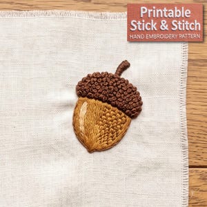 Può includere: Un disegno di ghianda ricamato su tessuto beige chiaro. La ghianda presenta un cappuccio testurizzato marrone scuro e una noce marrone dorato con cuciture dettagliate. Il testo "Printable Stick & Stitch HAND EMBROIDERY PATTERN" è visibile.