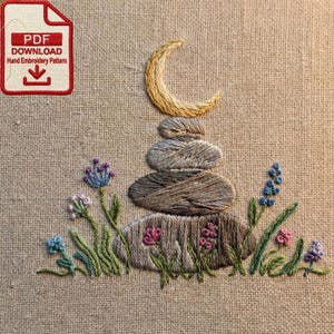 Pode incluir: Design bordado à mão com uma pilha de pedras, uma lua crescente e flores silvestres em tons de azul, rosa e verde. O design está em um tecido bege. Um ícone de download PDF vermelho está no canto superior esquerdo.